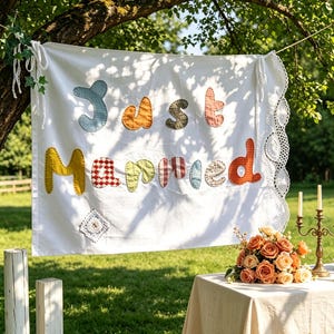 Op de afbeelding: Een wit stoffen spandoek met de tekst "Just Married" in kleurrijke, bedrukte applicatieletters. Het spandoek heeft een decoratieve kanten rand aan de rechterkant en is opgehangen met witte linten. Linksonder is een klein vierkant kanten lapje bevestigd.