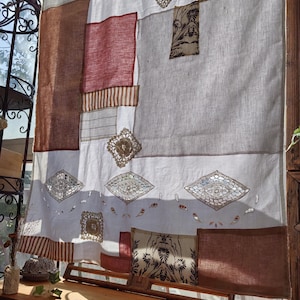 Pode incluir: Uma cortina patchwork com painéis de tecido em tons de castanho, vermelho e branco. A cortina apresenta padrões decorativos de diamantes e florais, criando uma estética boémia. A cortina está pendurada em frente a uma janela.