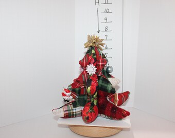 May include: A decorative Christmas tree made of red and green plaid fabric. The tree is adorned with a gold star, a white snowflake, a silver ornament, and a small Santa Claus figure. It sits on a wooden base.