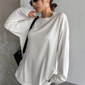 May include: A long-sleeve white crewneck shirt with a relaxed fit, paired with grey pants featuring the word SHINE printed on the leg. The outfit includes black sunglasses, large hoop earrings, and a black shoulder bag with a strap.