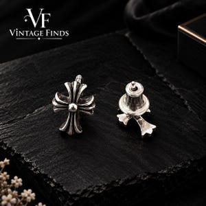 May include: A pair of silver stud earrings featuring a cross design with rounded, flared arms and a central circular detail. The earrings include a post and a standard butterfly clutch backing, displaying the text VF VINTAGE FINDS.