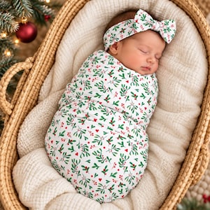 May include: A swaddled infant wearing a matching headband with a bow, both featuring a white fabric base printed with a pattern of green mistletoe leaves and small red berries, resting inside a woven basket lined with a soft cream blanket.