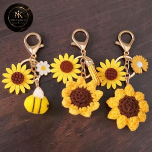 Puede incluir: Tres llaveros de metal dorado con dijes de girasol. El primero tiene un girasol de fieltro amarillo, una margarita blanca y una abeja amarilla y negra. El segundo tiene un girasol de fieltro y uno de ganchillo. El tercero tiene un girasol de ganchillo.