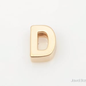 May include: A gold-colored metal letter "D" charm. The charm is in a serif font and has a three-dimensional appearance. The letter is set against a white background.