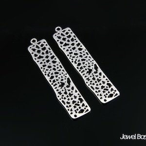 Pode incluir: Dois pingentes retangulares de filigrana prateada com um design delicado e intrincado. Os pingentes têm um furo na parte superior para serem presos a joias.