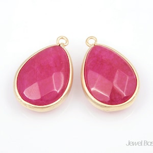 May include: Two pink teardrop-shaped gemstone pendants with gold-toned metal settings. The pendants are faceted and have a shiny finish.