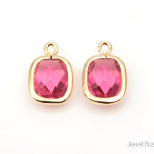 May include: Two gold-toned pendant charms with a square-shaped pink crystal in the center. The charms have a loop at the top for attaching to a necklace or bracelet.