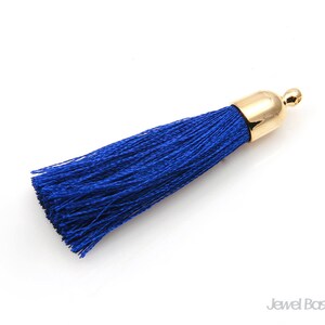 May include: A royal blue tassel with a gold-colored cap. The tassel is made of many strands of blue thread. The gold cap has a small loop at the top. The tassel is on a white background.