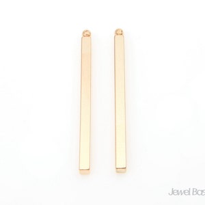 Puede incluir: Dos colgantes de barra de metal de color dorado con un bucle en la parte superior. Las barras son rectas y tienen un acabado liso y pulido.