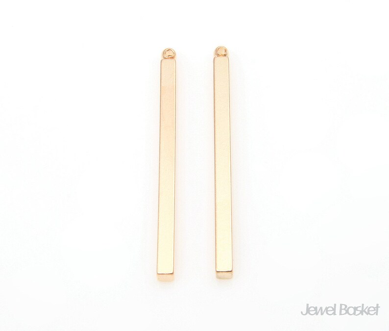 Puede incluir: Dos colgantes de barra de metal de color dorado con un bucle en la parte superior. Las barras son rectas y tienen un acabado liso y pulido.