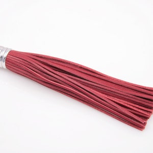 Puede incluir: Un borla de cuero rojo con una parte superior de metal plateado. La borla mide aproximadamente 15 cm de largo.