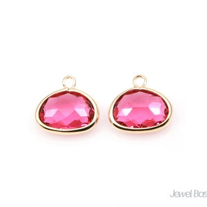 May include: Two gold-toned pendant charms featuring a pink faceted glass stone. The charms have a loop at the top for attaching to a necklace or bracelet.