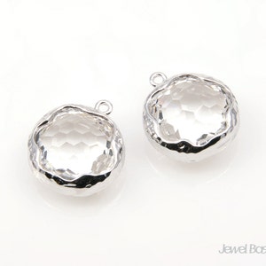 May include: Two silver-toned pendant charms with clear faceted glass stones. The stones are set in a hammered metal frame.