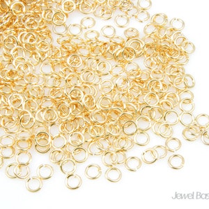 Puede incluir: Primer plano de un montón de pequeños anillos de salto dorados. Los anillos son redondos y tienen un acabado metálico brillante. Están dispersos sobre una superficie blanca, creando un patrón denso y texturizado. Ideal para la creación de joyas.