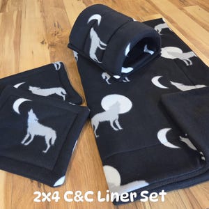May include: A black fleece pet cage liner set featuring a white wolf and moon pattern. The set includes a large rectangular mat and a smaller square mat, both with a soft texture. The text 2x4 C&C Liner Set is printed at the bottom.