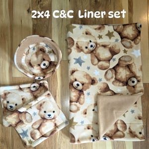 May include: A 2x4 C&C Liner set featuring a rectangular mat, a square pad, and a round bowl, all made of cream-colored fleece with a repeating pattern of brown teddy bears and gray and gold stars. The fabric has a soft, plush texture.