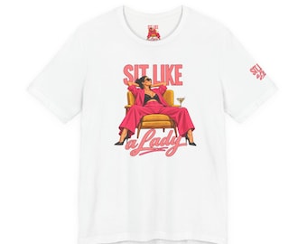 Könnte beinhalten: Ein weißes Kurzarm-T-Shirt mit einer Grafik einer Person im rosa Anzug, die in einem gelben Sessel mit einem Martiniglas sitzt. Der Text lautet SIT LIKE a Lady in Pink und Rot. Ein kleines Logo befindet sich am linken Ärmel und im Nackenbereich.