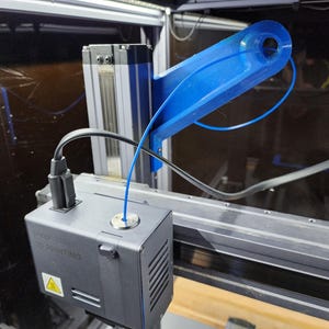May include: A gray 3D printer print head assembly featuring a blue filament guide arm and a black cable connection. The print head housing displays the text 3D PRINTING and a yellow warning symbol on its side, with a thin blue filament strand.