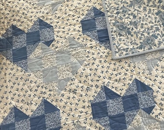 Op de afbeelding: Een doorgestikte deken met een patchworkpatroon van blauwe en crèmekleurige stoffen. Het ontwerp bevat hartvormige motieven bestaande uit effen blauwe en bloemrijke vierkanten, afgewisseld met een crèmekleurige stof bedrukt met kleine blauwe strikjes.