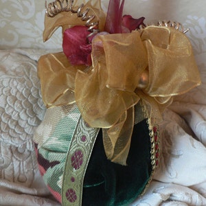 May include: A decorative ornament featuring a green velvet base, adorned with a gold ribbon and a patterned fabric. The ornament is embellished with red accents and topped with a gold bow and decorative elements.