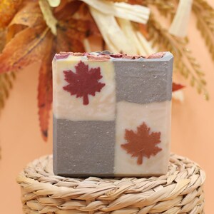May include: A square bar of soap featuring a four-quadrant design with alternating cream and gray sections. Two quadrants display a dark red maple leaf imprint, while the top edge is decorated with small, dried botanical pieces.