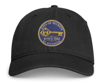 May include: A black baseball cap featuring a circular embroidered patch on the front. The patch has a blue background with a gold key graphic and the text CITY OF SECRETS SINCE 1565 ST. AUGUSTINE in white and gold lettering.