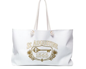 May include: A white canvas tote bag with two thick rope handles. The front features a gold and beige graphic with the text ST. AUGUSTINE, SINCE 1565, and CITY OF SECRETS, centered around an illustration of an ornate key inside a decorative shield.