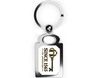 May include: A silver-toned metal keychain featuring a rectangular pendant with a white center insert. The insert displays a dark blue graphic of an ornate key and the text "SINCE 1565" and "ST. AUGUSTINE" in a vertical orientation.