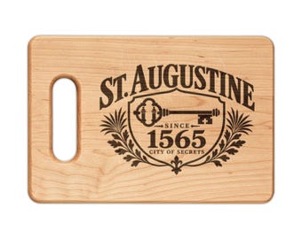 May include: A rectangular light-colored wooden cutting board with a handle cutout on the left side. The center features a dark brown laser-engraved design of a key, laurel leaves, and the text St. Augustine Since 1565 City of Secrets.