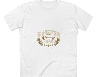 May include: A white short-sleeved t-shirt featuring a gold graphic on the chest. The design includes the text ST. AUGUSTINE, the year 1565, and the words CITY OF SECRETS, centered around an illustration of a key inside a shield emblem.