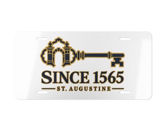 May include: A rectangular white license plate featuring a dark blue and gold graphic of an ornate key with an arched bow. Below the key, the text reads SINCE 1565 in large dark blue letters, followed by ST. AUGUSTINE in smaller gold letters.