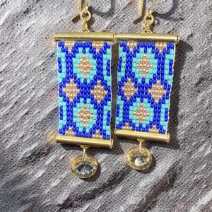 May include: A pair of gold-toned earrings with a rectangular beaded design in shades of blue, turquoise, and gold. Each earring has a small, clear, round charm dangling from the bottom.