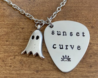 May include: A silver-colored necklace with a ghost charm and a guitar pick-shaped pendant. The pendant is stamped with the words "sunset curve" and a sun design. The necklace is on a wooden surface.
