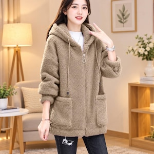 May include: A tan, long-sleeved, zip-up hoodie made of a textured, fuzzy fleece material. The garment features a hood with long drawstrings, a silver-toned circular zipper pull, and two large, square patch pockets on the front.