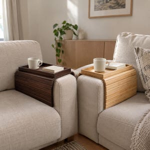 May include: Two flexible sofa arm trays, one in dark brown wood and one in light natural wood, each featuring a flat top surface. Each tray holds a white ceramic mug and a small rectangular book, resting securely over the arm of a beige sofa.