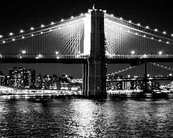 May include: A black and white photograph of the Brooklyn Bridge at night, featuring illuminated suspension cables and stone towers over the East River. The city skyline is visible in the background. The text Brooklyn Bridge, NYC is printed below.