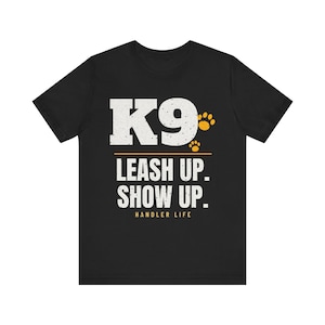 May include: A black short-sleeve t-shirt featuring the white text K9 with two small yellow paw prints. Below this, a yellow horizontal line separates the white text LEASH UP. SHOW UP. and the yellow text HANDLER LIFE at the bottom.