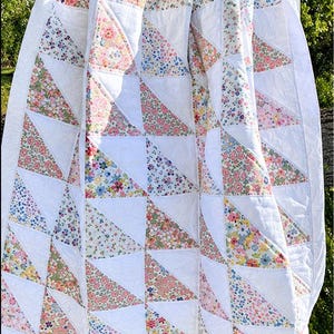 Op de afbeelding: Een quilt met een herhalend patroon van witte driehoeken en driehoeken met bloemenprint in roze, blauwe, gele en groene tinten. De stof is in een raster aan elkaar genaaid, wat een geometrisch ontwerp met een witte rand creëert.
