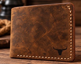Puede incluir: Una cartera bifold de cuero marrón con textura desgastada y costuras claras visibles en los bordes. La esquina inferior derecha presenta un pequeño gráfico negro en silueta de un cráneo de toro con cuernos.