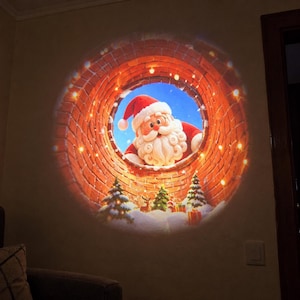 May include: A circular light projection on a wall displaying a festive scene of Santa Claus looking down from a brick chimney. The image features a blue sky, snow-covered ground, small evergreen trees, a reindeer, and wrapped gifts.