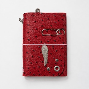May include: A red journal cover with a textured ostrich-like pattern, secured by a light pink elastic band. It features a silver-toned metal wing charm, a large silver paperclip, and two silver metal eyelets on the front cover.