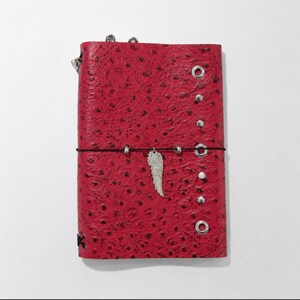 May include: A red journal cover with a textured ostrich-like pattern, featuring silver-toned metal grommets along the right edge, a black elastic closure band, and a silver-toned metal wing charm hanging from the center of the band.
