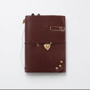 May include: A dark brown leather journal cover featuring a gold heart-shaped locket charm, a small gold star charm, and a gold heart-shaped paperclip. The cover includes three gold rivets at the bottom corner and a silver chain attached to the side.