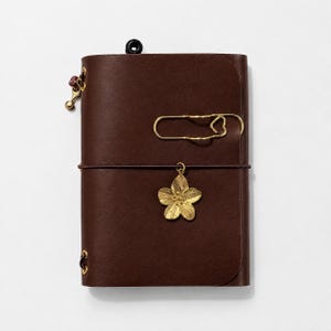 May include: A dark brown leather notebook cover featuring a gold-toned metal paperclip shaped like a heart and a gold-toned flower charm attached to a thin elastic band that wraps horizontally around the center of the cover.