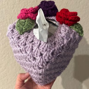 Könnte beinhalten: Ein gehäkelter Taschentuchbox-Bezug in helllila mit drei gehäkelten Blumen oben. Die Blumen sind rosa, rot und lila.