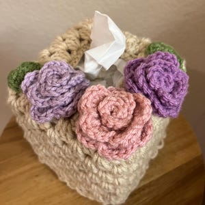 Könnte beinhalten: Ein gehäkelter Taschentuchbox-Bezug mit einer beigefarbenen Basis und drei gehäkelten Blumen in Rosa, Lila und Grün.