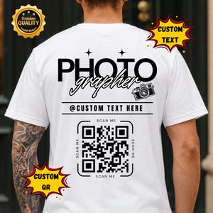 May include: A white t-shirt featuring black text that reads PHOTO grapher with a camera icon, the text @CUSTOM TEXT HERE, and a QR code surrounded by the words SCAN ME. The design includes two star icons above the word PHOTO.