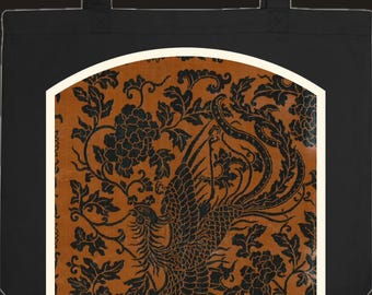 Può includere: Una borsa tote nera con una grafica rettangolare centrata e parte superiore ad arco. La grafica mostra un dettagliato motivo nero di una fenice ed elementi floreali su uno sfondo arancione bruciato, incorniciato da un bordo spesso color crema.