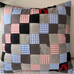 Peut inclure: Coussin carré en patchwork avec une variété de tissus en forme de grille. Les carrés comprennent du noir, du gris, du vichy rouge et blanc, du vichy bleu et blanc et des tissus gris unis. Le coussin a une bordure noire.