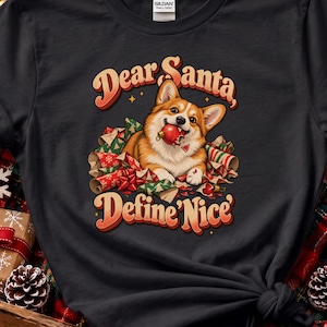 May include: A black t-shirt featuring a graphic of a Corgi dog holding a broken red Christmas ornament in its mouth, surrounded by torn wrapping paper. The text reads Dear Santa, Define Nice in a retro orange and red font style.